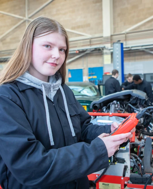 Student in a black overall holding a diagnostic tool in an automotive workshop, surrounded by mechanical equipment. Student in a black overall holding a diagnostic tool in an automotive workshop, surrounded by mechanical equipment.